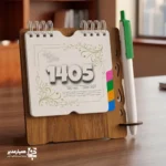 "📆 تقویم رومیزی کد 101 | انتخابی شیک برای میزهای حرفه‌ای تقویم رومیزی کد 101 با طراحی چوبی و ساختار جمع‌وجور، گزینه‌ای بسیار کاربردی برای میز کار در محیط‌های اداری، آموزشی و سازمانی است. این محصول با ظاهر گرم چوب و طراحی مینیمال، همزمان حس نظم، زیبایی و حرفه‌ای بودن را به میز شما می‌بخشد. بدنه این تقویم از MDF دورو با کیفیت بالا ساخته شده و ابعاد پایه آن 13×13.3 سانتی‌متر است که فضای کمی اشغال کرده و برای هر نوع میز کاری مناسب می‌باشد. برگه‌های تقویم با ابعاد 10.7×9.6 سانتی‌متر از کاغذ گلاسه 200 گرم تهیه شده‌اند که علاوه بر خوانایی بالا، از دوام و ماندگاری مناسبی برخوردار هستند. صحافی فنری، ورق‌زدن آسان و استفاده روزمره را بسیار راحت کرده است. ⭐ تقویم رومیزی کد 101 در رنگ‌بندی متنوع (چوب تیره، فندقی و طرح چوب طبیعی) تولید می‌شود تا با سلیقه‌ها و دکوراسیون‌های مختلف هماهنگ باشد. مهم‌ترین مزیت این محصول، قابلیت شخصی‌سازی اختصاصی آن است؛ امکان درج نام، لوگو یا اطلاعات سازمان، مدرسه یا برند این تقویم را به یک هدیه تبلیغاتی ماندگار و مؤثر تبدیل می‌کند. کد محصول: 101 جنس بدنه: MDF دورو ابعاد پایه: 13×13.3 سانتی‌متر ابعاد برگه: 10.7×9.6 سانتی‌متر کاغذ: گلاسه 200 گرم قابلیت شخصی‌سازی اختصاصی تقویم رومیزی کد 101؛ انتخابی هوشمندانه برای میز کار، هدیه سازمانی و تبلیغات ماندگار ✅ "