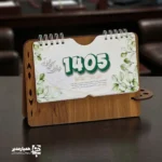 "📆 تقویم رومیزی کد 202 | انتخابی شیک برای میزهای حرفه‌ای تقویم رومیزی کد 202 با طراحی چوبی و ساختار جمع‌وجور، گزینه‌ای بسیار کاربردی برای میز کار در محیط‌های اداری، آموزشی و سازمانی است. این محصول با ظاهر گرم چوب و طراحی مینیمال، همزمان حس نظم، زیبایی و حرفه‌ای بودن را به میز شما می‌بخشد. بدنه این تقویم از MDF دورو با کیفیت بالا ساخته شده و ابعاد پایه آن 13*20 سانتی‌متر است که فضای کمی اشغال کرده و برای هر نوع میز کاری مناسب می‌باشد. برگه‌های تقویم با ابعاد 8.5*16 سانتی‌متر از کاغذ گلاسه 200 گرم تهیه شده‌اند که علاوه بر خوانایی بالا، از دوام و ماندگاری مناسبی برخوردار هستند. صحافی فنری، ورق‌زدن آسان و استفاده روزمره را بسیار راحت کرده است. ⭐ تقویم رومیزی کد 202 در رنگ‌3 رنگ متنوع (گردوجنگلی ،فندوقی،گردویی تیره ) تولید می‌شود تا با سلیقه‌ها و دکوراسیون‌های مختلف هماهنگ باشد. مهم‌ترین مزیت این محصول، قابلیت شخصی‌سازی اختصاصی آن است؛ امکان درج نام، لوگو یا اطلاعات سازمان، مدرسه یا برند این تقویم را به یک هدیه تبلیغاتی ماندگار و مؤثر تبدیل می‌کند. کد محصول: 202 جنس بدنه: MDF دورو ابعاد پایه13*20 سانتی‌متر ابعاد برگه:16*8.5سانتی‌متر کاغذ: گلاسه 200 گرم قابلیت شخصی‌سازی اختصاصی تقویم رومیزی کد 202؛ انتخابی هوشمندانه برای میز کار، هدیه سازمانی و تبلیغات ماندگار ✅ "