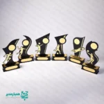 " تندیس ورزشی مدل قهرمان؛ نماد درخشش و پیروزی 🥇✨ تجلیل از تلاش‌های ورزشی دانش‌آموزان، یکی از موثرترین راه‌ها برای تقویت اعتمادبه‌نفس و ایجاد انگیزه در آن‌هاست. تندیس ورزشی چوبی مدل قهرمان، با طراحی پویا و نمادین، به‌خوبی حس حرکت و موفقیت را منتقل می‌کند. این تندیس‌ها به گونه‌ای طراحی شده‌اند که با قرارگیری در دکور اتاق یا بوفه افتخارات دانش‌آموز، همواره خاطره خوش پیروزی را برای او زنده نگه دارند. طراحی هوشمندانه و متریال باکیفیت 🎨 در ساخت این محصول از تکنولوژی برش لیزری دقیق استفاده شده است. بدنه اصلی از چوب MDF مشکی با ضخامت استاندارد و المان‌های ورزشی از پلکسی‌گلاس طلایی (آینه‌ای) ساخته شده‌اند. این ترکیب کنتراست رنگی زیبایی ایجاد کرده و باعث می‌شود تندیس در زیر نور سالن‌های ورزشی درخشش فوق‌العاده‌ای داشته باشد. تنوع رشته‌های ورزشی ⚽🏀🏐 یکی از بزرگترین مزایای مدل «قهرمان»، پوشش تمامی رشته‌های محبوب است. از جمله: فوتبال و فوتسال: با طرح‌های ضربه به توپ و دروازه‌بانی. بسکتبال و والیبال: نمایش فیگورهای تخصصی پرتاب و اسپک. دوومیدانی و آمادگی جسمانی: نماد سرعت و استقامت. هندبال و ورزش‌های رزمی: متناسب با نیاز مسابقات تخصصی. مشخصات فنی و ابعادی 📌 جنس بدنه: MDF درجه یک و پلکسی‌گلاس طلایی پایه: دارای پایه چوبی مستحکم (قابلیت نصب پلاک حکاکی شده) رنگ‌بندی: ترکیب مشکی و طلایی (بسیار رسمی و لوکس) کاربرد: المپیادهای ورزشی، جشن‌های پایان سال، مسابقات محلی 💡 پیشنهاد برای مدارس: پایه پهن این تندیس این امکان را به شما می‌دهد تا نام دانش‌آموز، نام مدرسه و سال تحصیلی را روی یک لیبل طلایی چاپ کرده و در قسمت جلوی پایه بچسبانید تا هدیه کاملاً اختصاصی و ماندگار شود. "